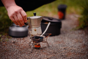 Moka Pot in hand, over fire burner, brewing coffee outdoors.