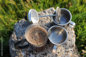French Press coffee maker in nature with two steel cups on a rock.