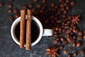 An overview of a white coffee cup filled with black coffee. Two cinnamon sticks rest on top of the cup, surrounded by scattered coffee beans and star anise on a dark surface.