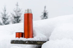 A red thermos standing in the snow with its lid off, resting on a wooden ledge. Snowy trees are faintly visible in the background.