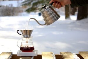A hand pouring hot water from a silver gooseneck kettle into a coffee dripper placed on a glass carafe. The setup is outdoors on a snowy surface, with steam rising from the pour.