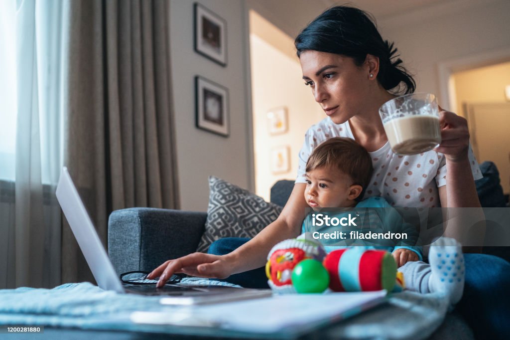 A working mother with a laptop, a coffee nearby, and her infant in her lap.
